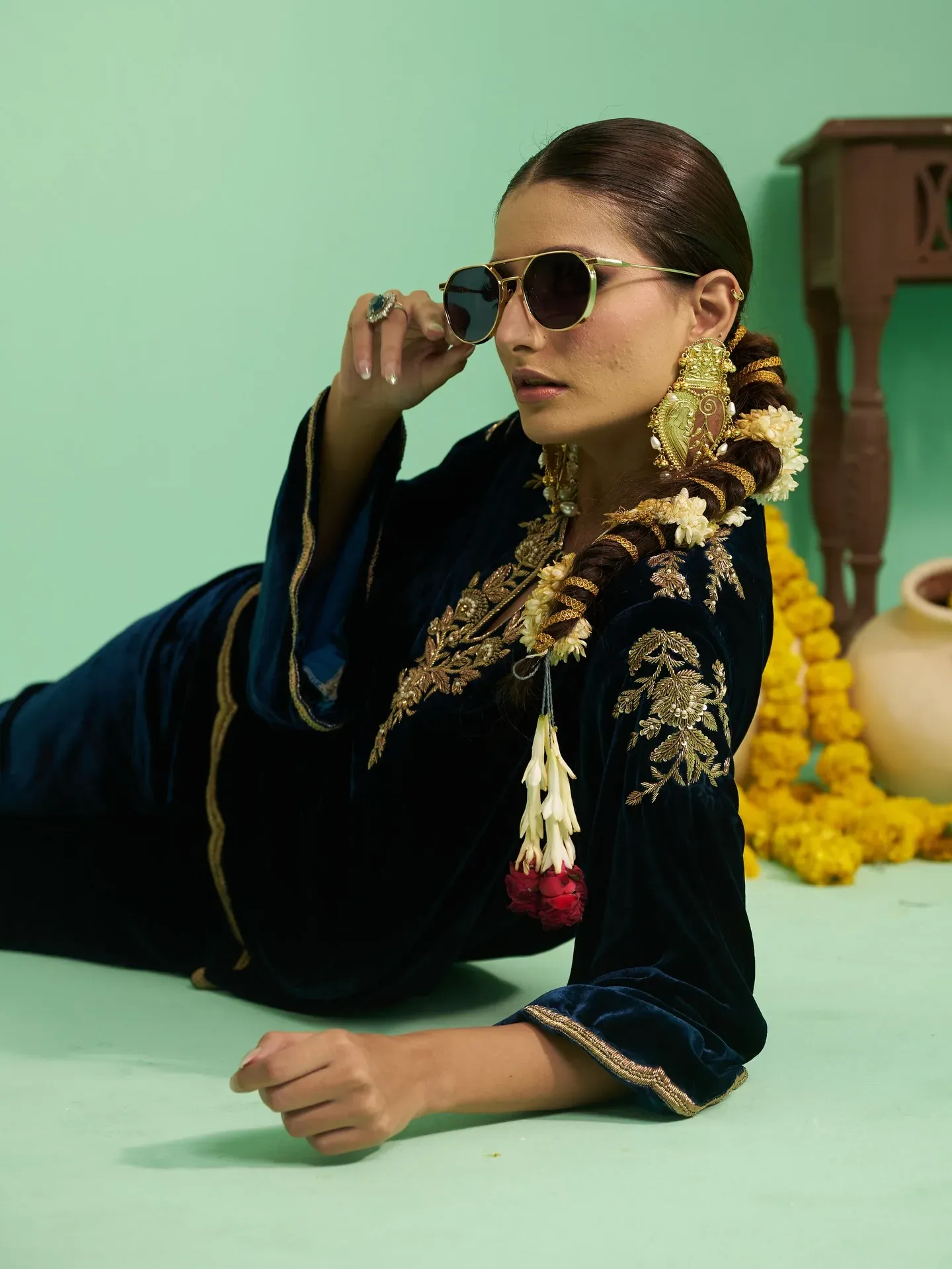 Model reclining in a navy velvet kurta with gold embroidery and aviator sunglasses, on a mint backdrop