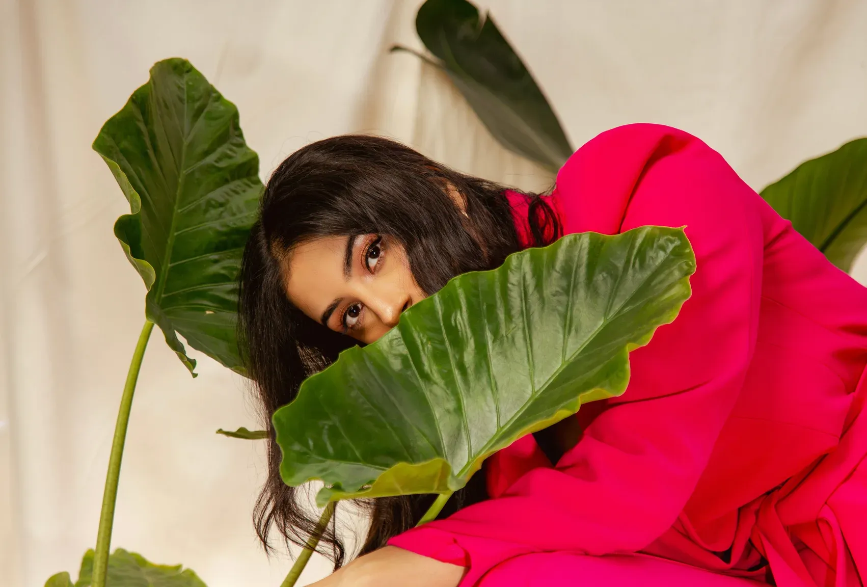 Model in fuchsia, framed by large green leaves against a draped ivory backdrop