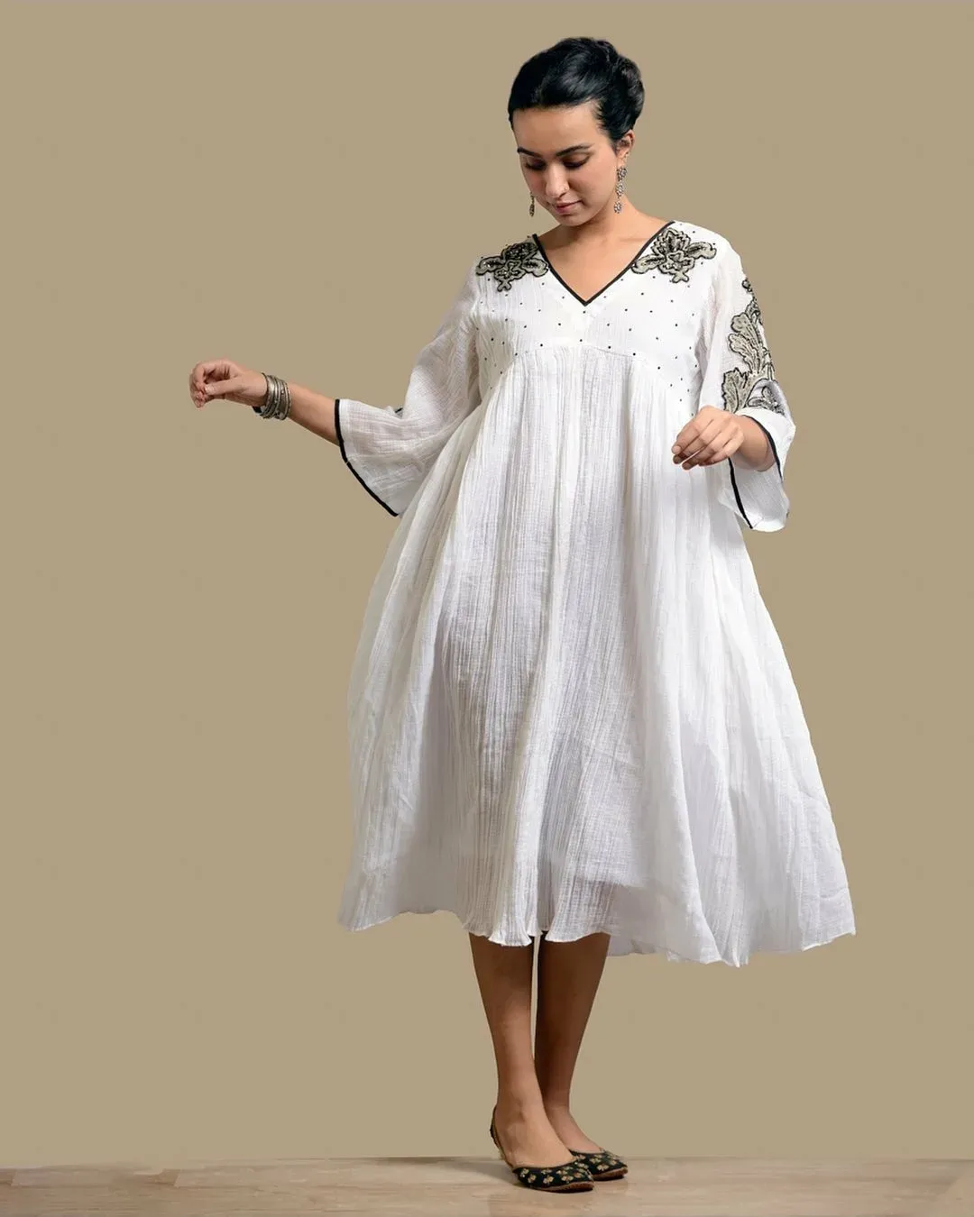 Model in a white crinkle-cotton kaftan dress with dark floral embroidery, against a beige studio backdrop