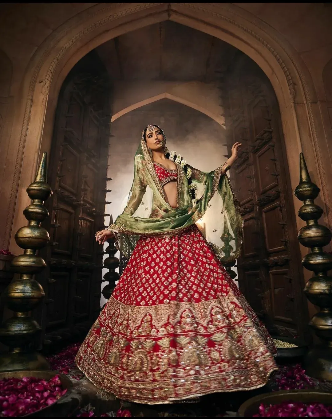 Model in a red and gold hand-embroidered lehenga with a green organza dupatta, framed by a carved arch and brass finials