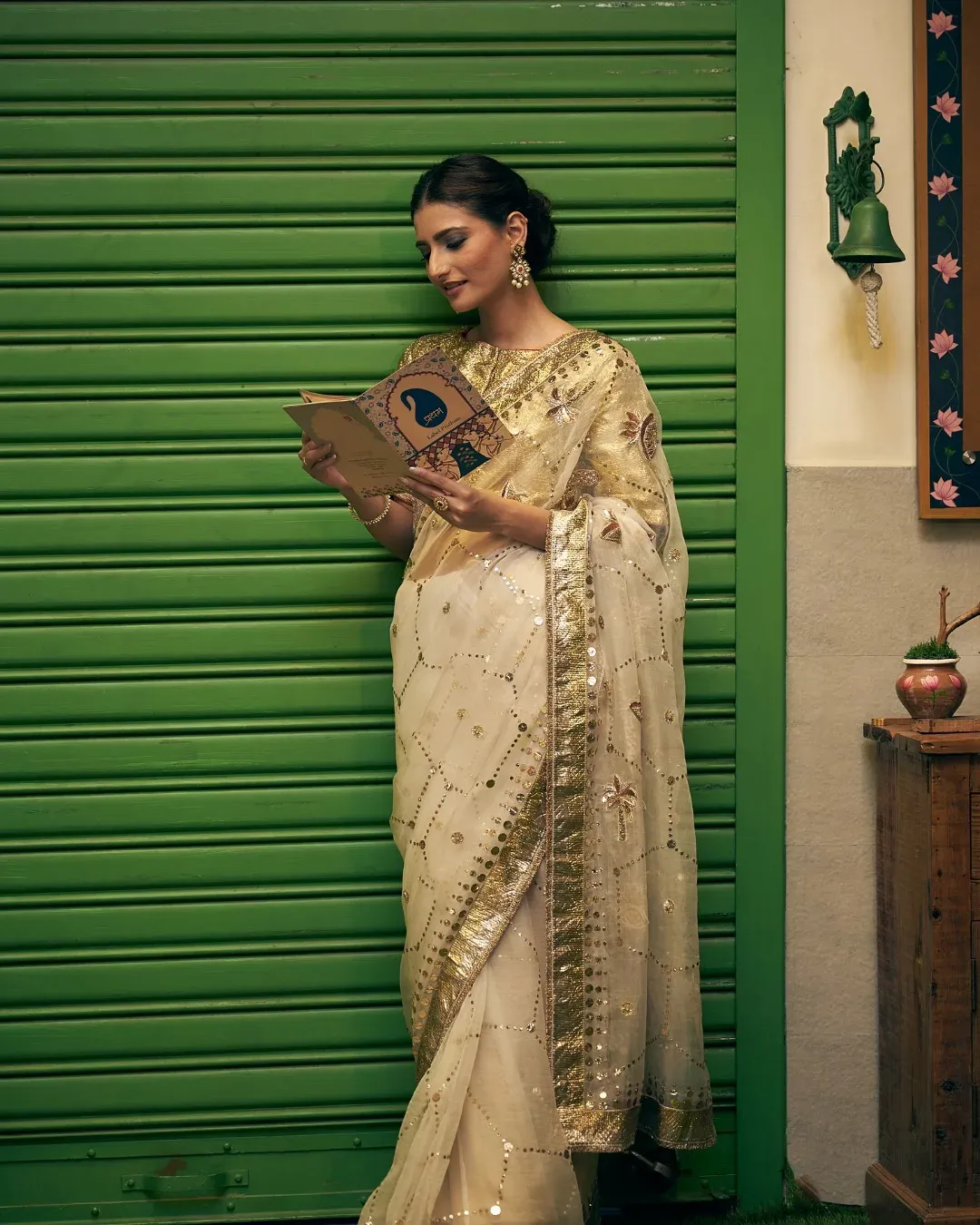 Model in an ivory embroidered saree holding a book, against a green rolling shutter