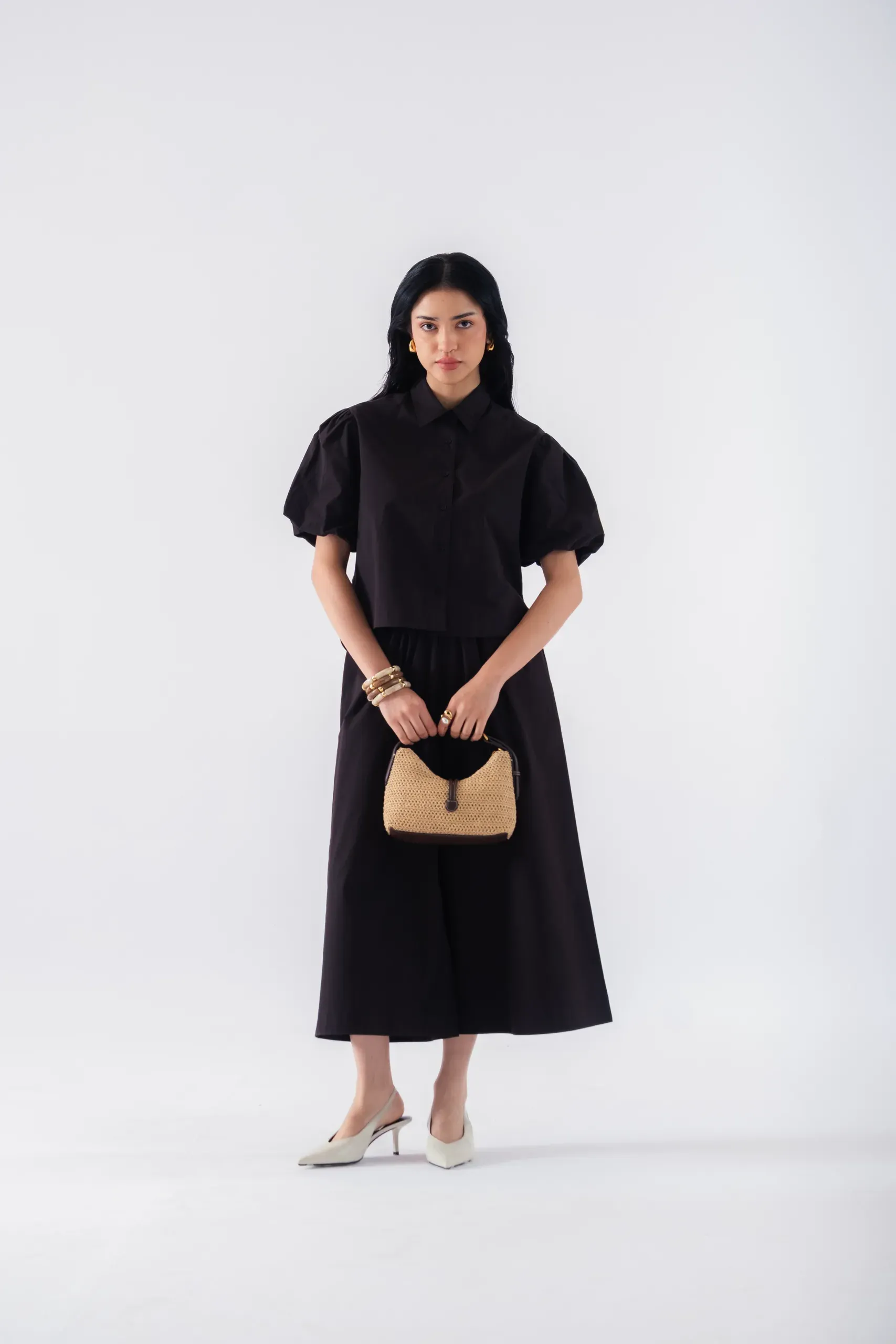Model in a black puff-sleeve shirt and wide culottes holding a woven raffia bag, on a white backdrop
