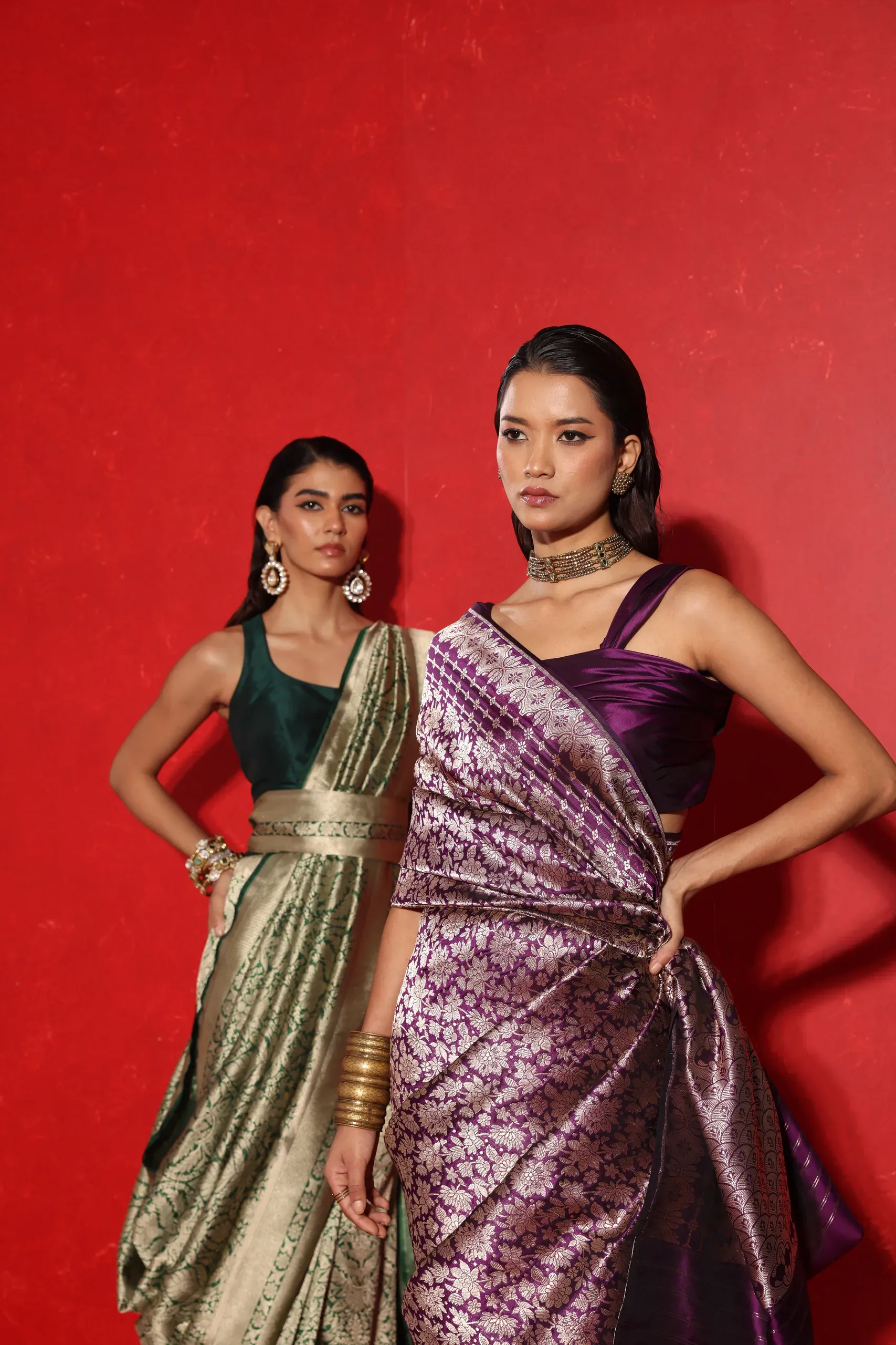 Two models in green and purple brocade sarees, posed against a red backdrop
