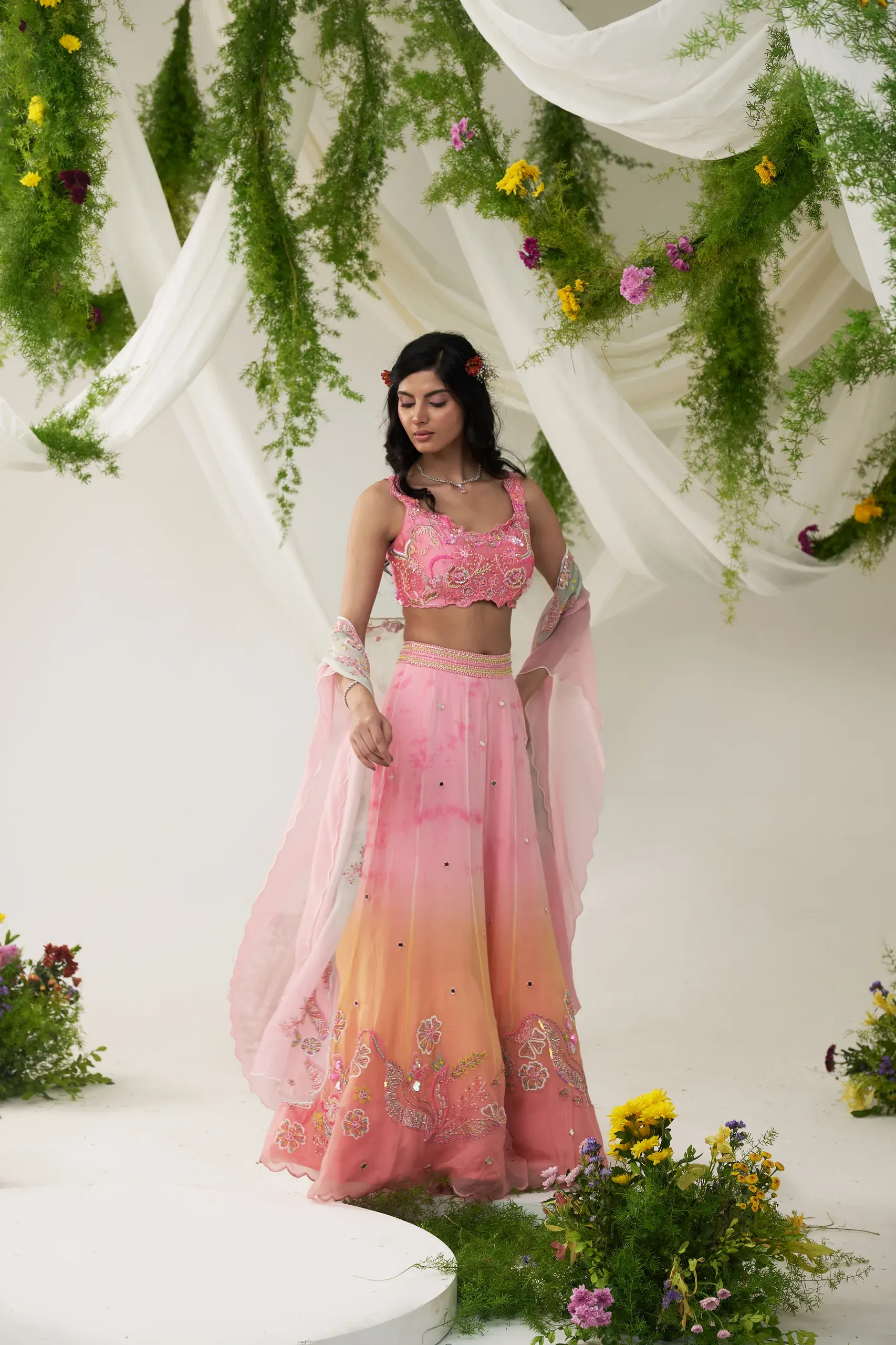 Model in a pink-to-peach ombré lehenga with mirrorwork, on a white set dressed with drapes and flowers