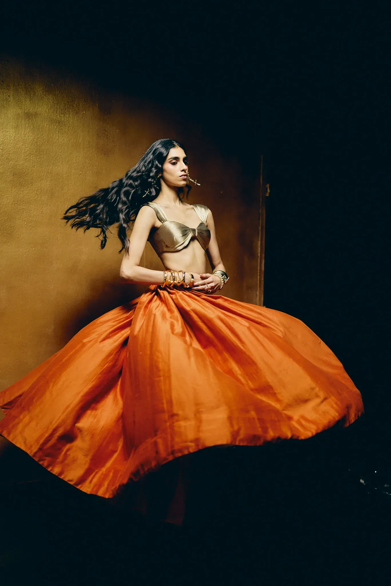Model mid-twirl in a full rust-orange skirt and metallic bralette against a gold backdrop