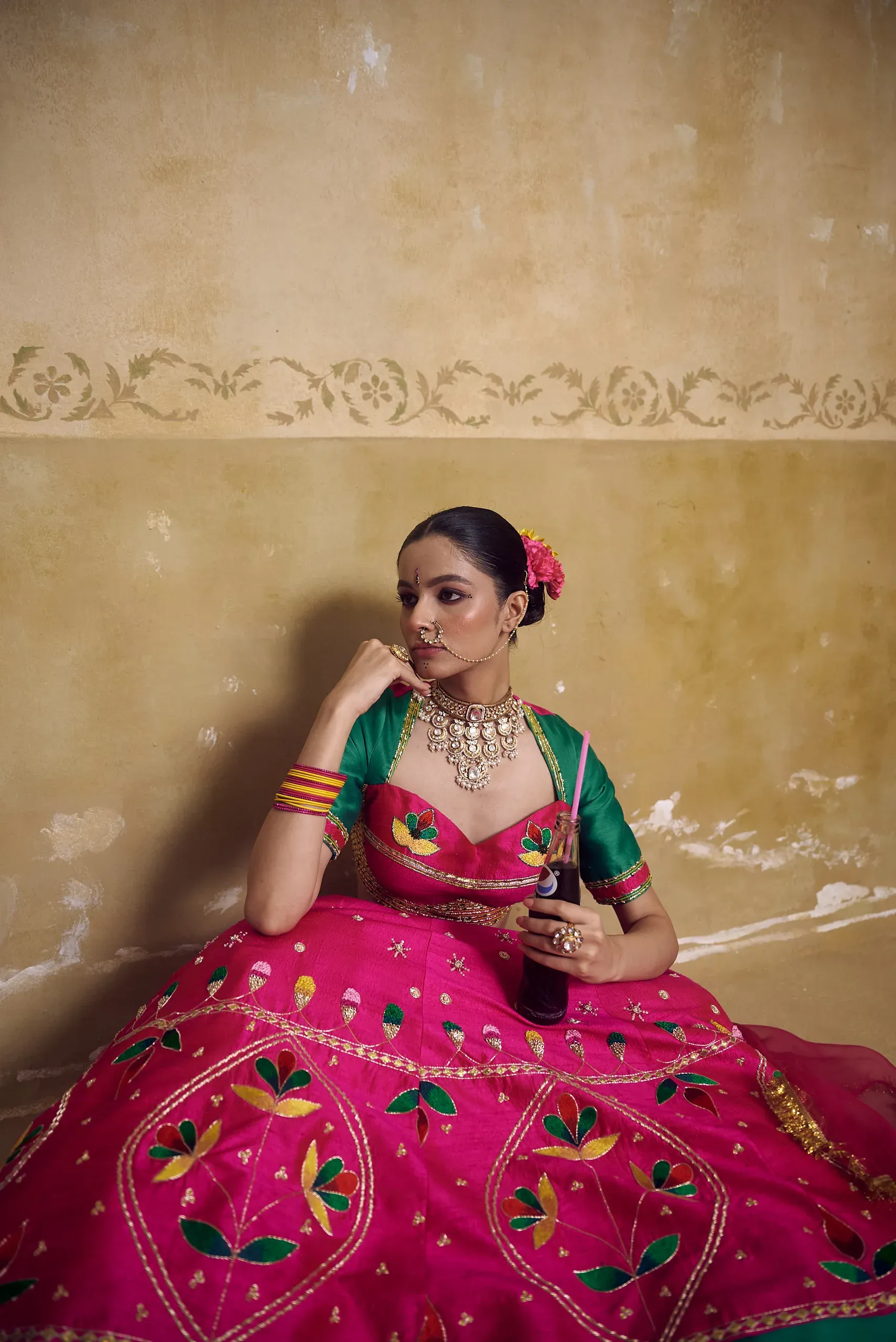Model in a fuchsia embroidered lehenga with a green blouse, seated against an ochre wall