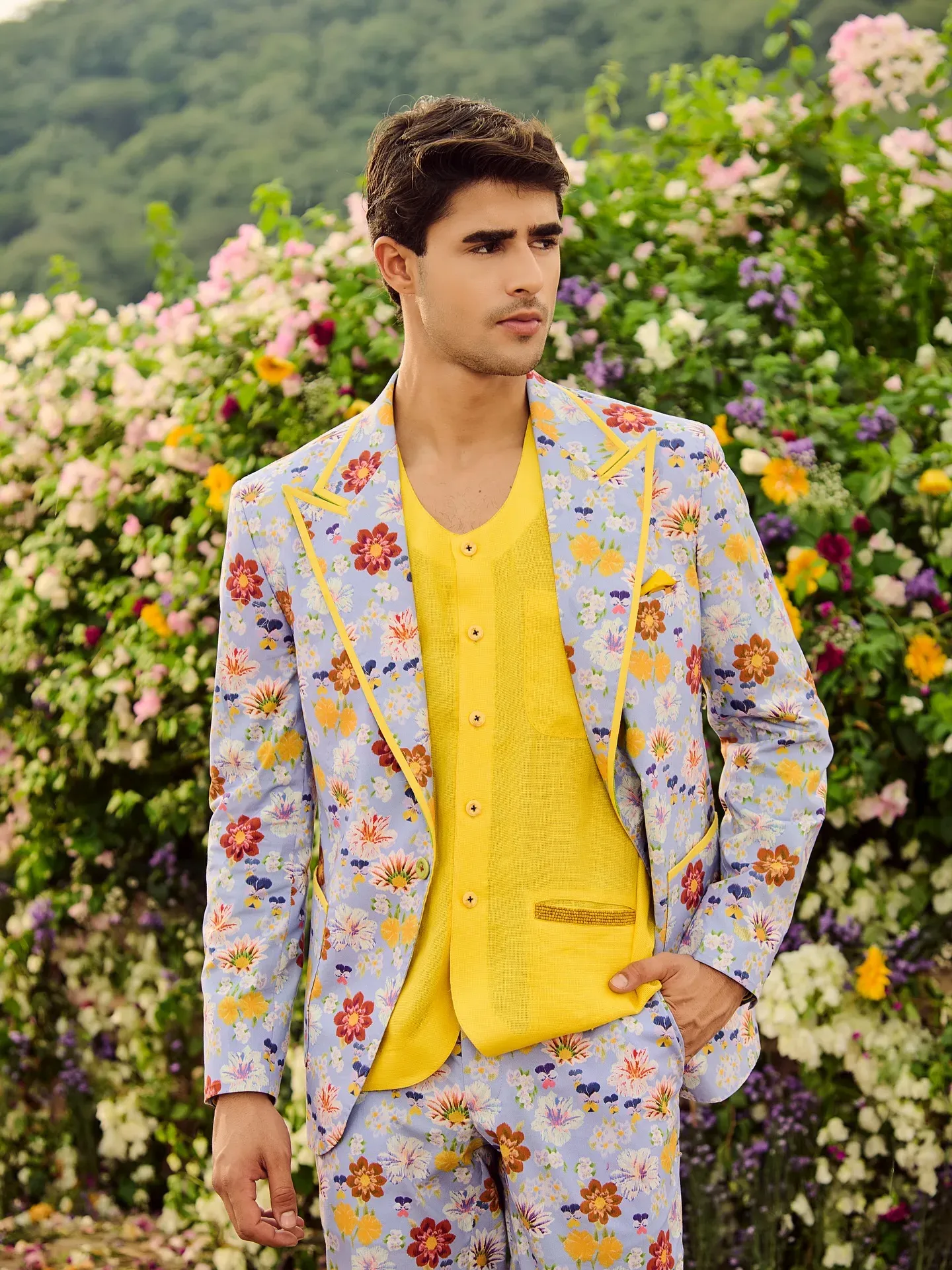 Model in a pastel floral-print suit over a yellow vest, standing before a flowering hedge
